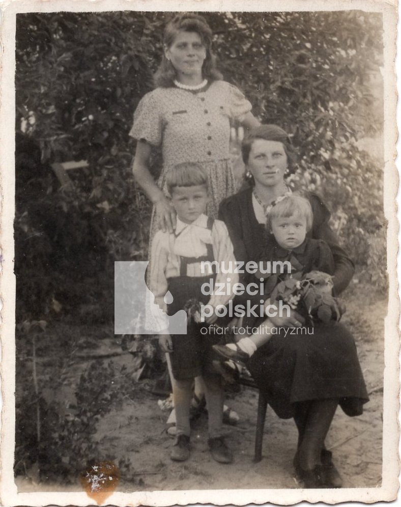 Fotografia rodzinna. Na pierwszym planie stoi kobieta Bronisława Suś i chłopiec Jan Śliwiński. Na krześle siedzi kobieta Władysława Śliwińska, która na kolanach trzyma córkę Krystynę. W tle ogród.