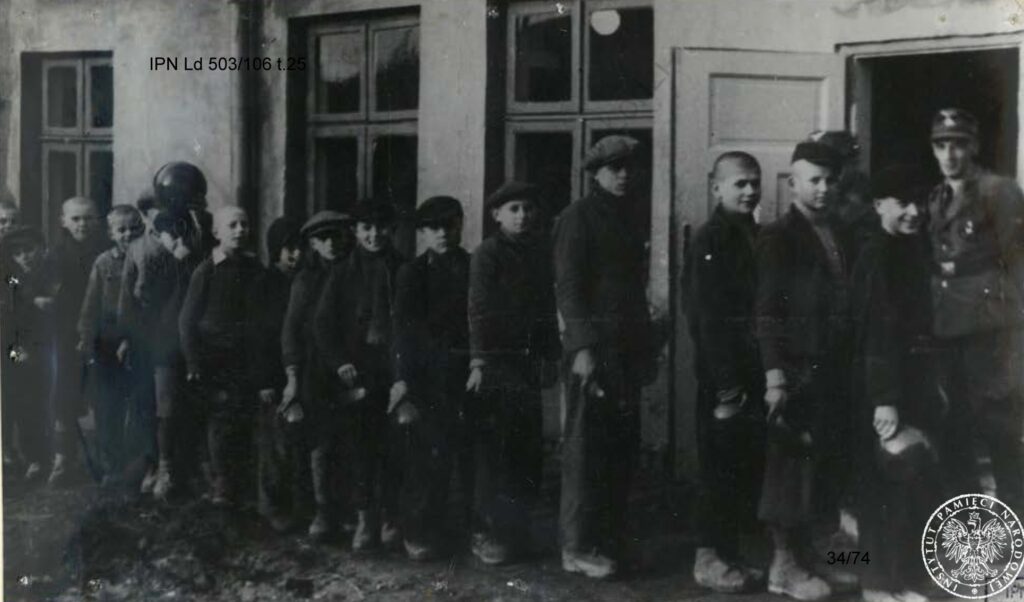 Kilkunastu chłopców ustawionych przed budynkiem, czeka na posiłek. W rękach trzymają miski. W otwartych drzwiach budynku stoi wachman.