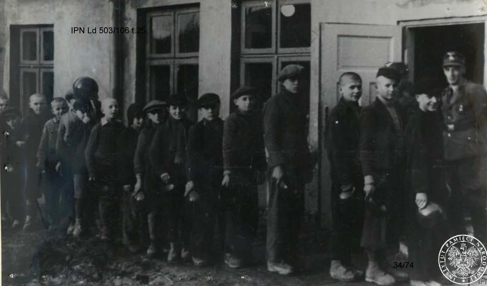 Kilkunastu chłopców ustawionych przed budynkiem, czeka na posiłek. W rękach trzymają miski. W otwartych drzwiach budynku stoi wachman.