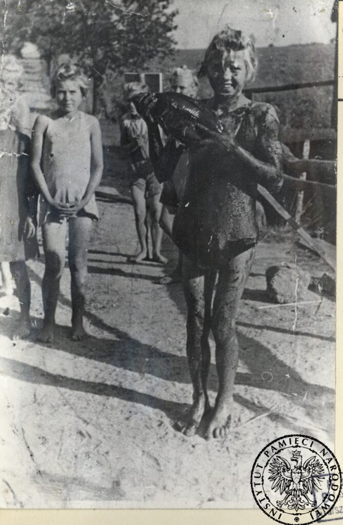 Na pierwszym planie uśmiechnięta dziewczynka, ubrudzona torfem, trzymająca w ręku rybę. Zdjęcie wykonane latem, na moście przy stawach. Na drugim planie inne więźniarki.