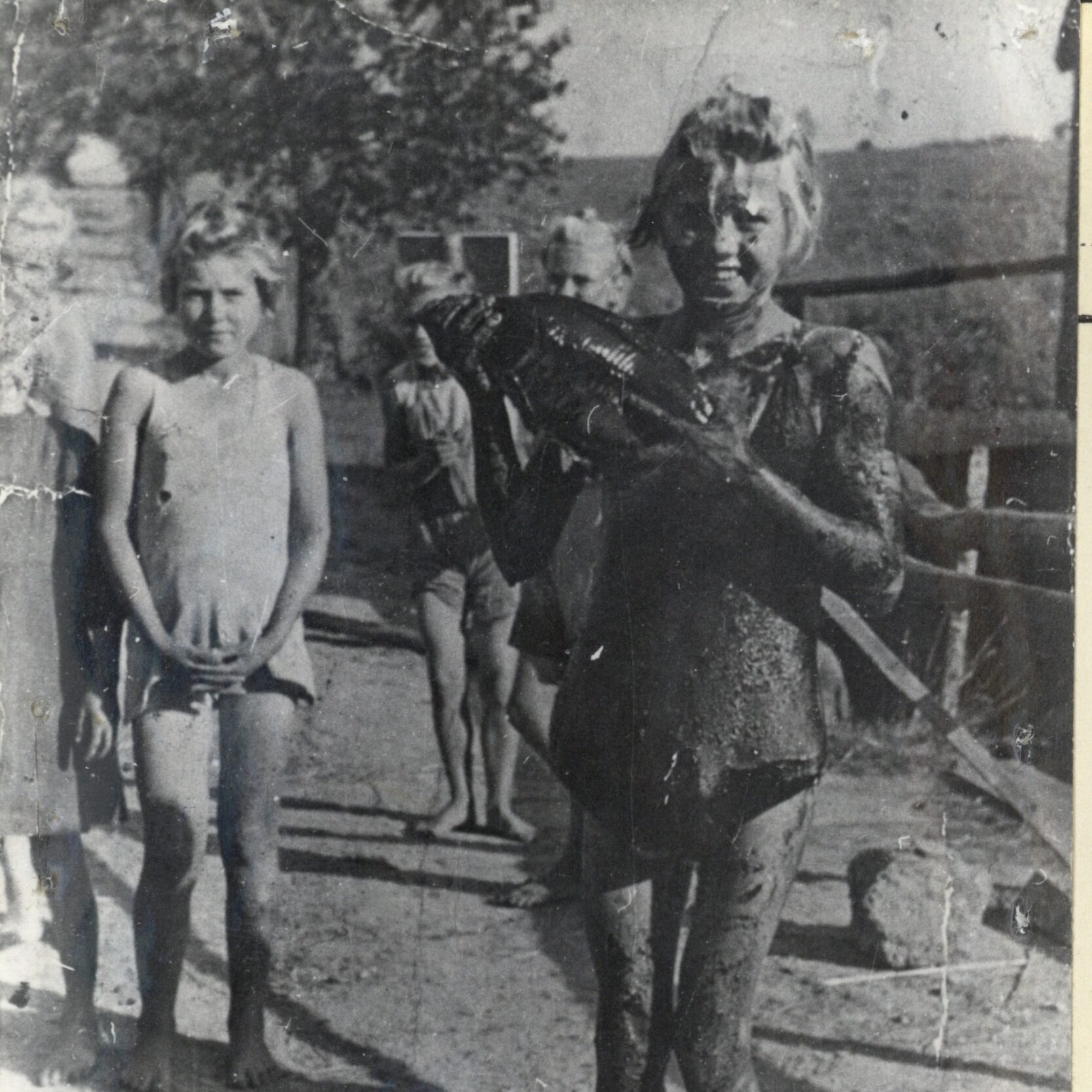 Na pierwszym planie uśmiechnięta dziewczynka, ubrudzona torfem, trzymająca w ręku rybę. Zdjęcie wykonane latem, na moście przy stawach. Na drugim planie inne więźniarki.